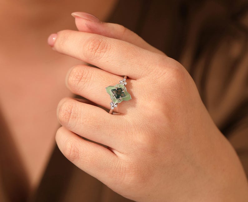 Puede incluir: Un anillo de plata con un dise&ntilde;o de tr&eacute;bol de cuatro hojas, con una piedra preciosa verde claro en el centro y peque&ntilde;as piedras claras. El anillo se lleva en una mano, mostrando sus delicados detalles y su elegante estilo.