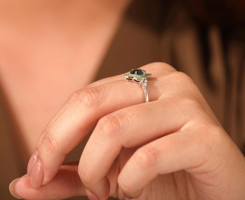 Puede incluir: Un anillo de plata con una piedra preciosa verde oscuro en el centro, rodeada de piedras blancas m&aacute;s peque&ntilde;as. El anillo tiene un dise&ntilde;o floral y se lleva en un dedo.
