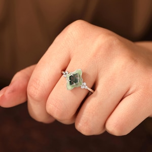 Puede incluir: Un anillo de plata con una piedra verde claro en forma de tr&eacute;bol de cuatro hojas con un centro verde oscuro. El anillo tiene peque&ntilde;as piedras de acento transparentes en la banda y alrededor de la piedra principal.