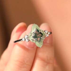 Puede incluir: Un anillo de plata con una piedra verde claro de forma irregular con aspecto de musgo. La piedra est&aacute; engastada en un engaste de esmalte verde claro con peque&ntilde;as piedras de acento transparentes. El anillo est&aacute; sostenido por una mano.