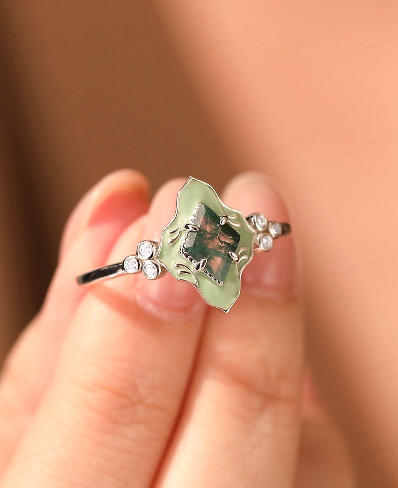 Puede incluir: Un anillo de plata con una piedra verde claro de forma irregular con aspecto de musgo. La piedra est&aacute; engastada en un engaste de esmalte verde claro con peque&ntilde;as piedras de acento transparentes. El anillo est&aacute; sostenido por una mano.