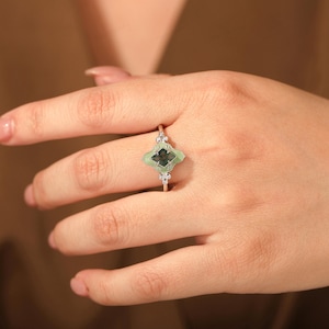 Puede incluir: Anillo plateado con una piedra verde en forma de tr&eacute;bol de cuatro hojas con un centro oscuro. El anillo tiene peque&ntilde;as piedras de acento transparentes a cada lado de la piedra principal. El anillo se muestra en una mano.