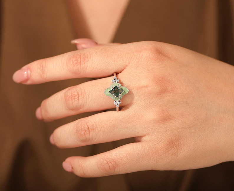 Puede incluir: Anillo plateado con una piedra verde en forma de tr&eacute;bol de cuatro hojas con un centro oscuro. El anillo tiene peque&ntilde;as piedras de acento transparentes a cada lado de la piedra principal. El anillo se muestra en una mano.