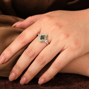 Puede incluir: Anillo plateado con una piedra preciosa verde claro de forma cuadrada y centro oscuro. Peque&ntilde;as piedras claras de acento flanquean la piedra principal. El anillo est&aacute; en un dedo. El dise&ntilde;o del anillo recuerda a un tr&eacute;bol de cuatro hojas.