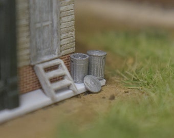 Puede incluir: Un modelo en miniatura de un edificio con una puerta blanca y escalones, dos pequeños cubos de basura plateados y una zona de césped.