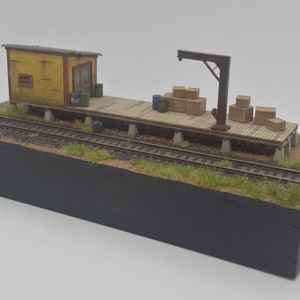 Puede incluir: Una escena de ferrocarril modelo con un edificio amarillo, un muelle de carga, una grúa y cajas. La escena está ambientada en una base de madera con hierba y tierra.