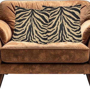 May include: Two square throw cushions featuring a bold black and beige tiger stripe pattern. The cushions are positioned side by side, displaying a consistent animal-inspired print with dark vertical stripes against a light tan background.