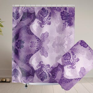 May include: A purple shower curtain and bath mat set with a floral watercolour design. The set features shades of purple and white. The curtain hangs on a white rail, and the mat is folded.