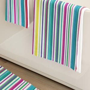 Pode incluir: Um conjunto de toalhas de banho e um tapete de banho a condizer com um padrão vibrante de riscas verticais em tons de magenta, azul-petróleo, roxo, amarelo e branco. O tecido tem uma textura macia com linhas consistentes e repetitivas.