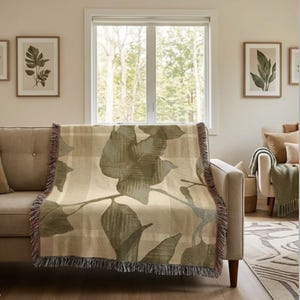May include: A woven throw blanket featuring a large-scale botanical leaf pattern in shades of sage green and olive layered over a neutral beige and cream plaid background, finished with decorative fringed edges on the top and bottom sides.
