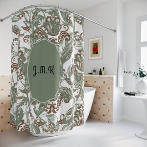 Puede incluir: Cortina de ducha blanca con un estampado floral verde y marrón. La cortina presenta un panel central verde oliva con las iniciales "J.M.K" en escritura negra. El diseño incluye hojas estilizadas y elementos decorativos.