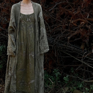 Puede incluir: Un vestido largo de lino verde fluido con una chaqueta a juego de manga larga. El vestido tiene un corpiño fruncido y una falda fluida. La chaqueta tiene un simple frente abierto y mangas largas. La persona está de pie frente a una pila de ramas.