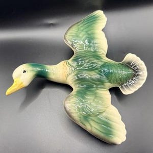 May include: A ceramic wall hanging in the shape of a flying duck. The duck features a cream-colored head and body with vibrant green feathers on its wings and back. It has a bright yellow beak and a small black eye, with sculpted wing details.