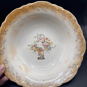 May include: A round ceramic bowl with a scalloped edge featuring a cream-colored base and a wide, light orange border. The rim is decorated with a grey floral pattern, and the center displays a colorful illustration of a flower-filled basket.