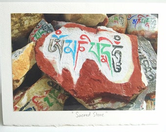 Op de afbeelding: Een foto van verschillende stenen, waarvan sommige beschilderd zijn met kleurrijke tekst. De centrale steen is rood met witte verf en blauwe, rode en groene tekst. De afbeelding heeft de titel "Sacred Stone".