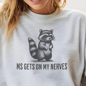 May include: A light gray t-shirt featuring a detailed illustration of a raccoon standing upright. The raccoon is black and gray with a striped tail. Below the raccoon is the text "MS GETS ON MY NERVES" in a sans-serif font.