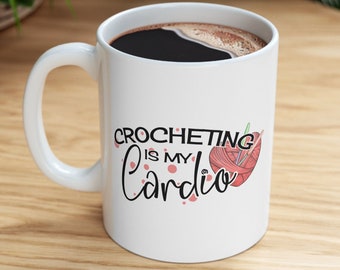 May include: White ceramic coffee mug with a black handle. The mug has a pink and white yarn ball with knitting needles and the text "Crocheting is my Cardio" printed on it.