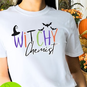 May include: White t-shirt with the text "Witchy Chemist" in a colorful, playful font, featuring a witch hat and bats. The shirt is a Halloween-themed design, perfect for a spooky season.