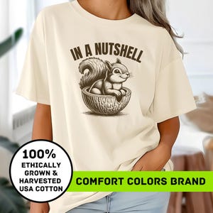 May include: A beige t-shirt with the text "In a Nutshell" and a cartoon squirrel holding a nut. The shirt also has the text "100% Ethically Grown & Harvested USA Cotton" and "Comfort Colors Brand".