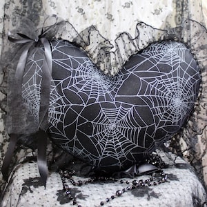 May include: A heart-shaped black pillow with a white spiderweb design. The pillow is trimmed with black lace and a black ribbon. A black beaded necklace is draped nearby. The pillow is on a lace-covered surface.