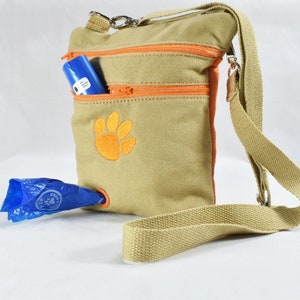 Op de afbeelding: Een beige canvas-hondenpoepzakdispenser met een oranje voetafdruk en twee oranje ritsvakken. De dispenser heeft een blauwe plastic zakdispenser en een blauwe plastic zak die uit de dispenser hangt. De zakdispenser heeft een verstelbare beige band.