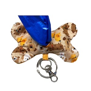 Op de afbeelding: Een bruine, botvormige hondendrollen-zakdispenser met een blauwe zak eraan en een zilveren sleutelhanger. Het bot is bedekt met een patroon van cartoonhonden.