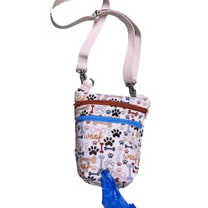 Op de afbeelding: Een witte canvas hondenpoepzakdispenser met een pootprint en "woof" patroon. De zakdispenser heeft een witte band en een blauwe plastic zak die aan de onderkant hangt.