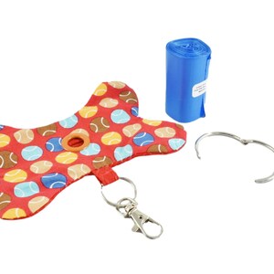 Op de afbeelding: Een rode, botvormige hondendrollen-zakdispenser met een kleurrijk tennisbalpatroon. Hij heeft een klein gaatje om zakken uit te geven en een metalen ring om aan een riem te bevestigen. Een rol blauwe plastic zakken en een metalen ring zijn ook inbegrepen.
