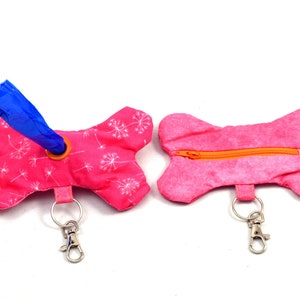 Op de afbeelding: Twee roze, botvormige hondenpoepzakjeshouders met een wit paardenbloemmotief. De ene houder heeft een blauwe zakdispenser en de andere een ritssluiting. Beide houders hebben zilveren sleutelhangers met een clip.