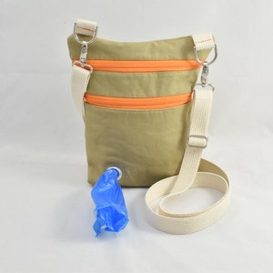 Op de afbeelding: Een beige canvas tas met een oranje rits en een witte band. De tas heeft een klein gaatje aan de onderkant voor een blauwe plastic zak.