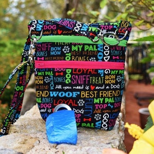 Op de afbeelding: Een zwarte crossbodytas met hondenthema met een kleurrijke print met woorden als "SNIFF", "WOOF" en "BONES". Het heeft een groene rits, een roze rits en een blauwe zakjesdispenser. De tas heeft een bijpassende riem en zilveren metalen ringen.