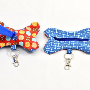 Op de afbeelding: Twee hondenbotvormige poepzakhouders. Eén is rood met een tennisbalpatroon en een blauwe poepzakdispenser. De andere is blauw met een botpatroon en een ritszak. Beide hebben een zilveren sleutelhanger.