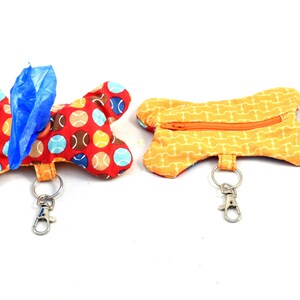 Op de afbeelding: Twee hondenbotvormige stoffen zakjes. Eén is rood met tennisbalpatronen en bevat een blauwe zak. De andere is geel met een botpatroon en een rits. Beide hebben sleutelringen.