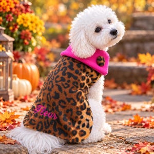 Könnte beinhalten: Ein weißer Hund trägt einen braun-schwarzen Fleece-Mantel mit Leopardenmuster, einem leuchtend pinken Kragen und einem einzelnen Knopf mit Leopardenmuster. Der Name Fleur ist in pinker Schreibschrift auf der Seite des Mantels eingestickt.