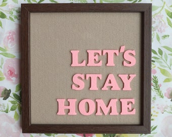 May include: A brown framed sign with the words "Let's Stay Home" in pink felt letters.