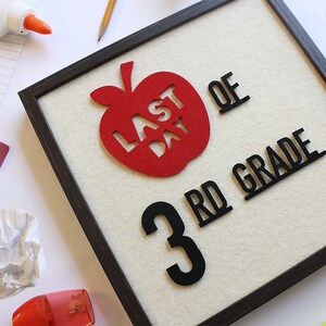 Puede incluir: Un cartel de fieltro enmarcado con una forma de manzana roja con las palabras "&Uacute;ltimo D&iacute;a" recortadas. Debajo de la manzana hay un n&uacute;mero negro "3" y las palabras "3er Grado" en negro.