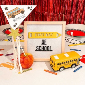 Puede incluir: Un cartel blanco con las palabras "First Day of School" en letras negras. Una forma de l&aacute;piz amarillo est&aacute; por encima de las palabras. Un autob&uacute;s escolar de juguete amarillo est&aacute; en primer plano. Una manzana roja y l&aacute;pices de colores est&aacute;n sobre la mesa.