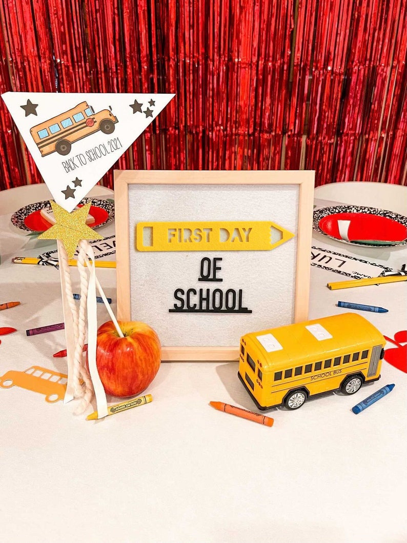 Puede incluir: Un cartel blanco con las palabras "First Day of School" en letras negras. Una forma de l&aacute;piz amarillo est&aacute; por encima de las palabras. Un autob&uacute;s escolar de juguete amarillo est&aacute; en primer plano. Una manzana roja y l&aacute;pices de colores est&aacute;n sobre la mesa.