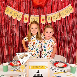 Puede incluir: Dos ni&ntilde;os con pijamas a juego con estampado de alfabeto se paran frente a una cortina de papel de aluminio roja con un globo rojo en forma de manzana. Una mesa est&aacute; preparada con un letrero que dice "Primer d&iacute;a de escuela" y un juguete de autob&uacute;s escolar.