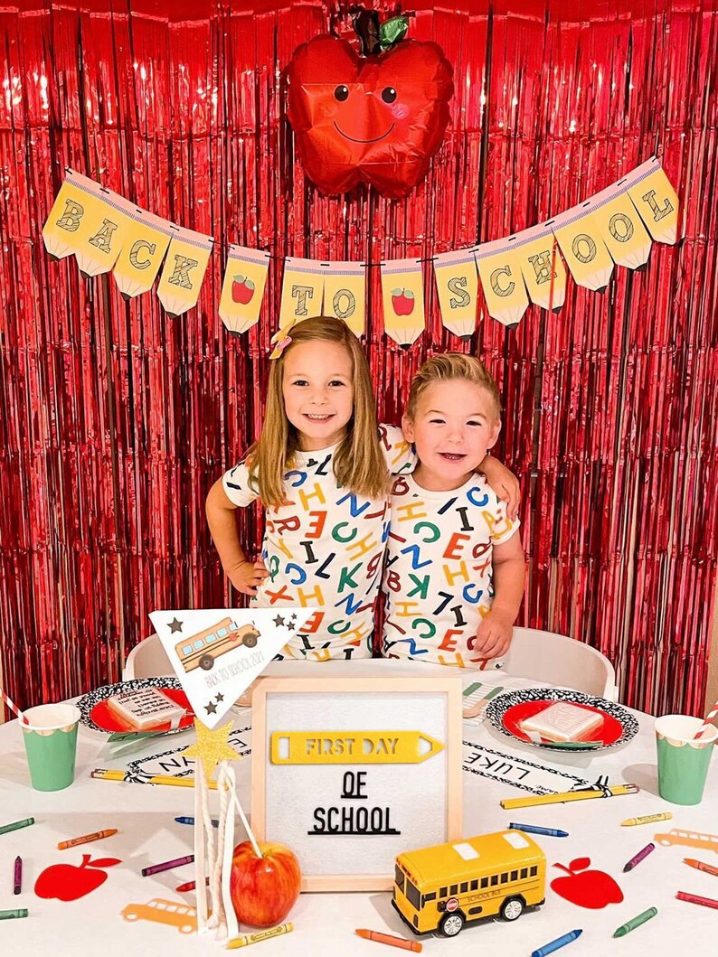Puede incluir: Dos ni&ntilde;os con pijamas a juego con estampado de alfabeto se paran frente a una cortina de papel de aluminio roja con un globo rojo en forma de manzana. Una mesa est&aacute; preparada con un letrero que dice "Primer d&iacute;a de escuela" y un juguete de autob&uacute;s escolar.