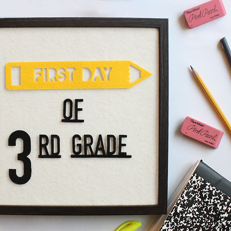 Puede incluir: Un tablero de fieltro enmarcado con las palabras "First Day of 3rd Grade" en letras de fieltro negro y amarillo. El tablero est&aacute; rodeado por un marco negro y est&aacute; sentado sobre una superficie blanca.