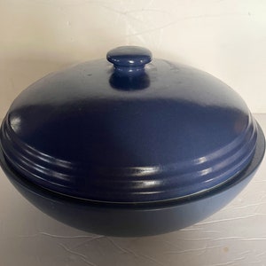May include: A navy blue ceramic casserole dish with a matching lid. The lid has a rounded knob and a series of concentric ridges. The dish has a rounded base and a slightly flared rim. The overall design is simple and functional.
