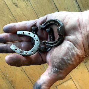 May include: Three weathered metal horseshoes, two complete and one partial, are held in a hand. The horseshoes are a dark, aged metal with visible wear and tear. The hand is covered in dark smudges, suggesting manual labor.