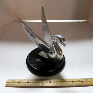 May include: Silver swan hood ornament with black base. The swan has its wings spread and its head turned to the side.