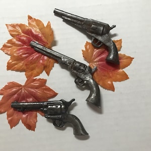Könnte beinhalten: Drei antike silberne Spielzeugpistolen auf weißem Hintergrund mit orangefarbenen und braunen Herbstblättern.