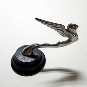 Puede incluir: Adorno de capó plateado que representa una figura alada en vuelo. La figura está montada sobre una base negra escalonada. El adorno tiene un diseño aerodinámico, con alas detalladas y líneas fluidas.