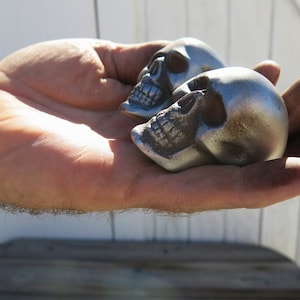 May include: Two silver-coloured skull figurines with detailed facial features are held in a hand. The skulls have a metallic sheen and are positioned close together. The background is blurred, suggesting an outdoor setting.