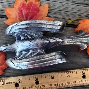 May include: A silver-colored vintage car hood ornament in the shape of a stylized bird with outstretched wings. The ornament is placed on a wooden surface with colorful autumn leaves and a ruler. The ruler shows measurements in inches.