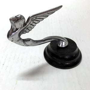 May include: A silver-toned art deco hood ornament depicting a winged figure in flight. The figure is mounted on a round, dark wooden base. The wings are outstretched, and the figure is gracefully posed.