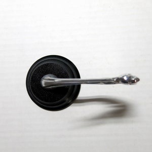 Puede incluir: Un tapón de botella negro con un asa plateada. El asa tiene un diseño decorativo en el extremo. El tapón está hecho de un material negro texturizado.